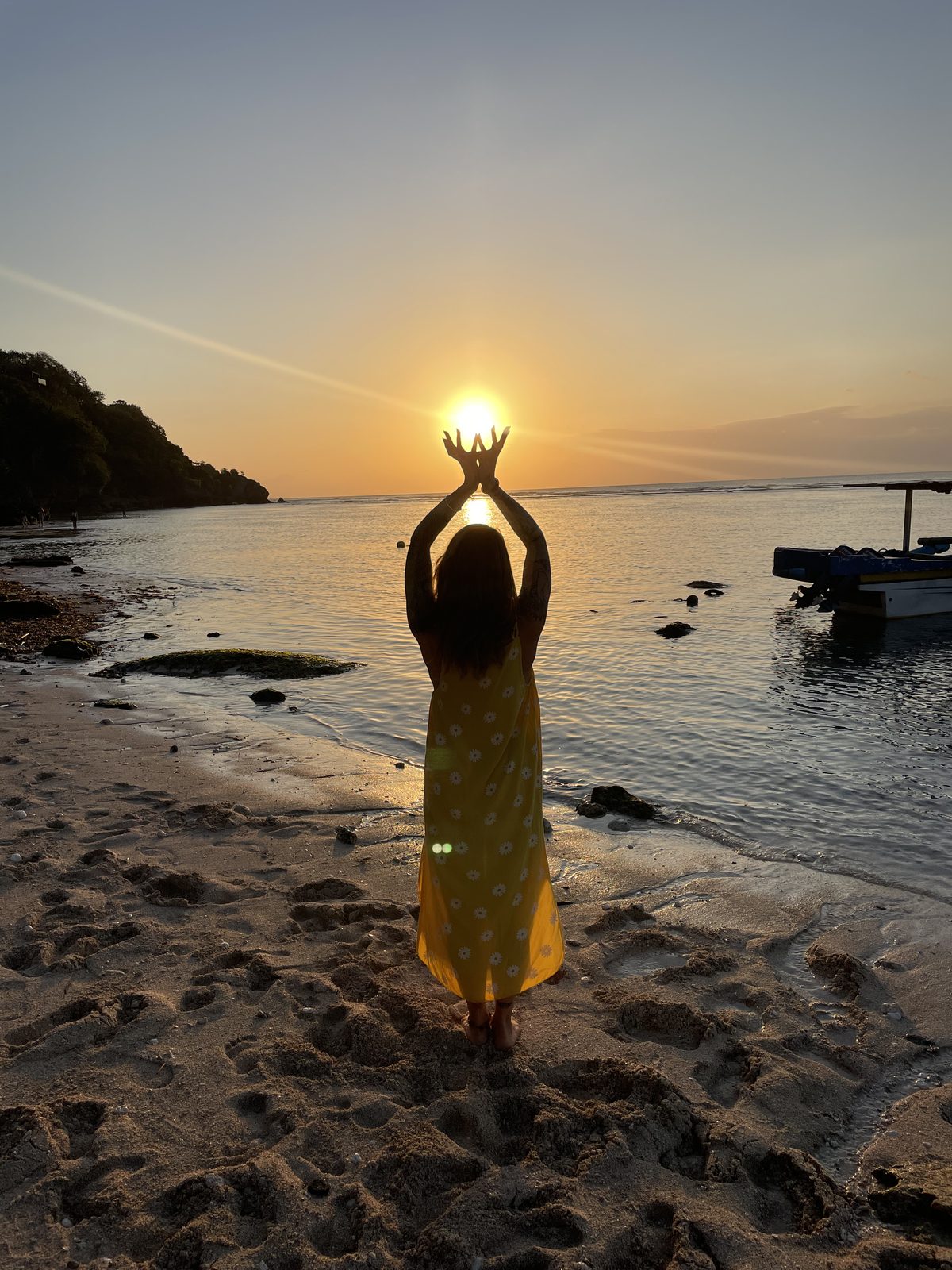 Cassandre standing on a beach at sunset, arms raised, framing the sun between her hands — taken in Bali
